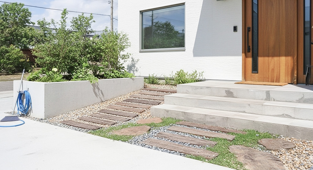 北欧ナチュラルスタイルの玄関アプローチ、枕木と砂利、グランドカバーを組み合わせた自然な小道、白い外壁と木製ドアのモダンな住宅